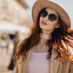 Good-looking european woman in black sunglasses plays with curly ginger hair. Outdoor photo of carefree red-haired female model in elegant hat walking down the street..